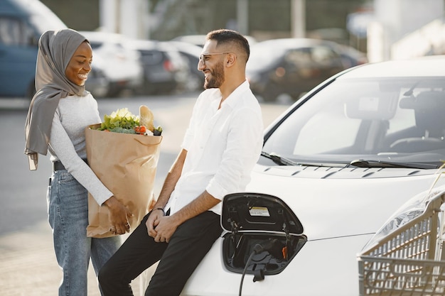 Pareja árabe africana se encuentra con comestibles cerca del coche eléctrico. Carga de coche eléctrico en la gasolinera eléctrica.