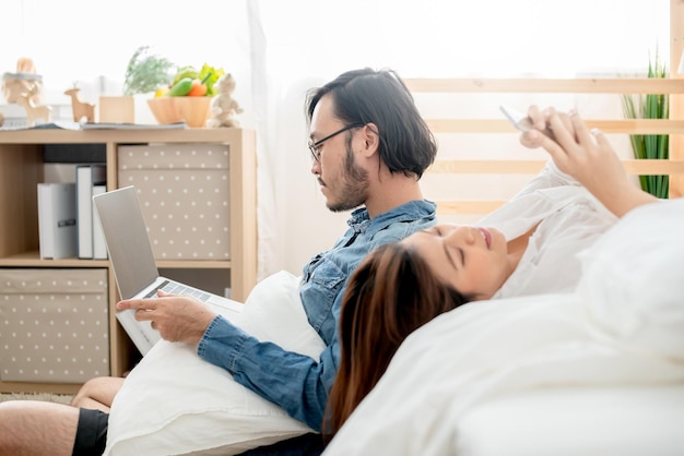 Pareja asiática amante de la actividad casual en la mano del dormitorio portátil y fondo interior del dormitorio del teléfono inteligente