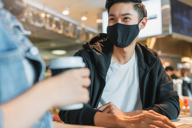 Una pareja asiática feliz usa protección contra virus con mascarilla en una cafetería disfruta de una buena conversación Un hombre y una mujer jóvenes en un restaurante mirando la computadora con pantalla táctil se ríen y sonríen juntos