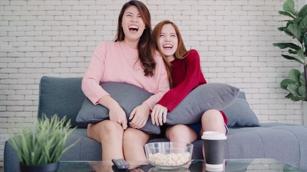 Pareja asiática lesbiana viendo la televisión reír y comiendo palomitas de maíz en la sala de estar en casa, dulce pareja disfruta