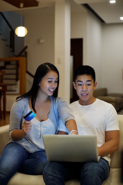 Pareja asiática sentada con laptop y tarjeta de crédito en el sofá y mirando la pantalla