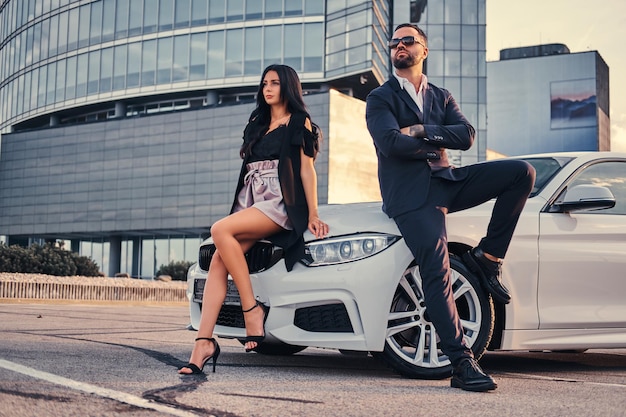 Pareja atractiva bien vestida apoyada en un coche de lujo al aire libre contra el rascacielos.