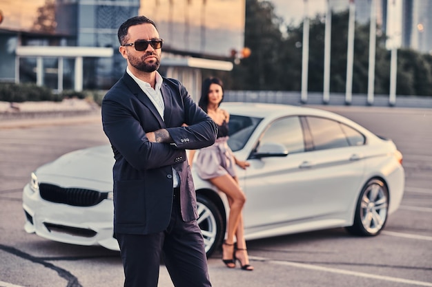 Pareja atractiva bien vestida apoyada en un coche de lujo al aire libre contra el rascacielos.