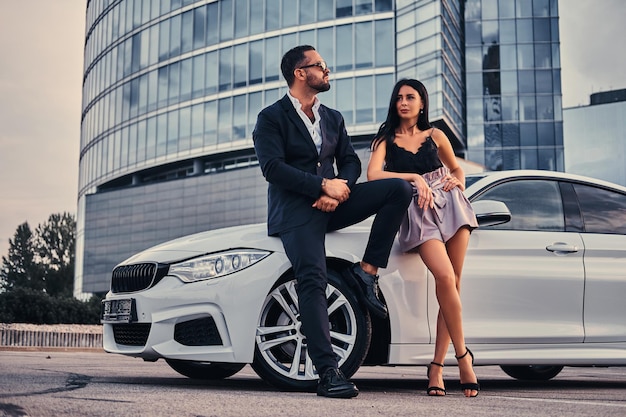 Pareja atractiva bien vestida apoyada en un coche de lujo al aire libre contra el rascacielos.