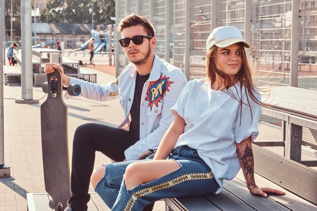 Pareja atractiva de jóvenes hipsters vestidos de moda con patinetas sentados en un banco en el complejo deportivo de la ciudad en un día soleado, con tonos cálidos.