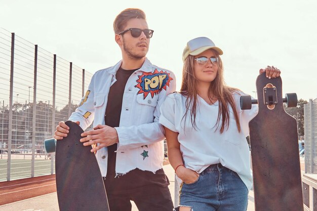 Pareja atractiva de jóvenes hipsters vestidos de moda posando con patinetas en el complejo deportivo de la ciudad en un día soleado, con tonos cálidos.