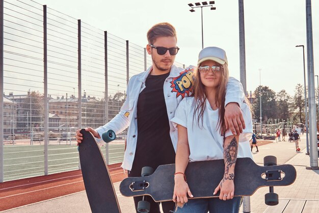 Pareja atractiva de jóvenes hipsters vestidos de moda posando con patinetas en el complejo deportivo de la ciudad en un día soleado, con tonos cálidos.