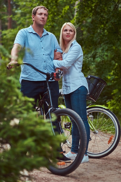 Una pareja atractiva de una mujer rubia y un hombre vestidos con ropa informal, se abrazan cerca de una bicicleta en un parque.