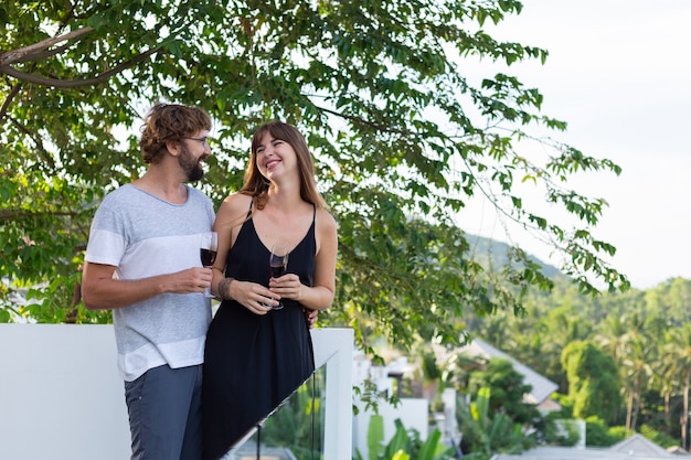 Pareja bebiendo vino en el balcón con vista de palmeras tópicas.