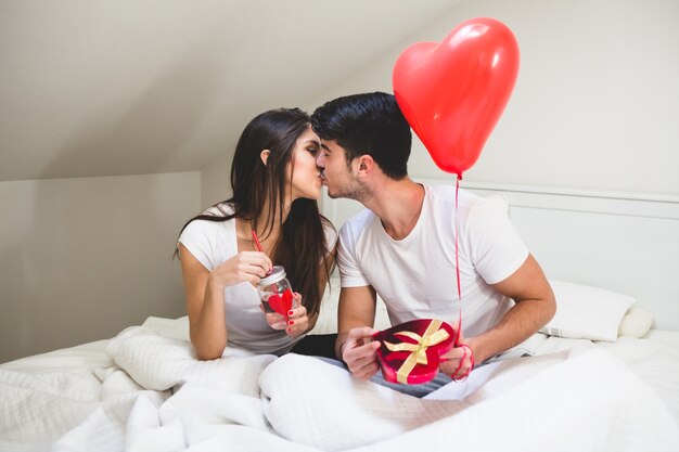 Pareja besándose mientras el sujeta un regalo y un globo y ella un vaso con agua