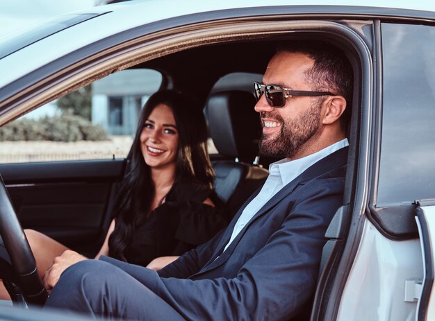 Una pareja bien vestida, un hombre barbudo sonriente y una mujer hermosa sentada en los asientos delanteros en un auto de lujo.