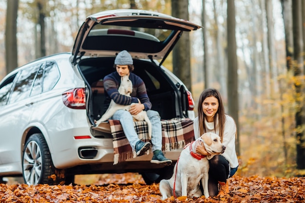 Pareja caminando con perros en el bosque de otoño, propietarios con labrador dorado relajándose cerca del coche.