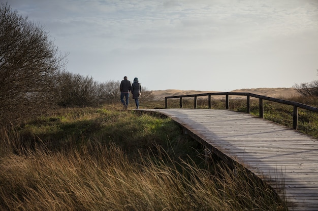 Pareja caminando sobre un puente de madera rodeado por un campo y colinas bajo la luz del sol