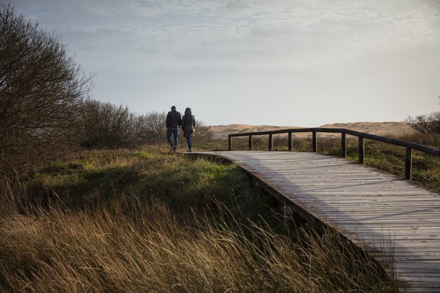 Pareja caminando sobre un puente de madera rodeado por un campo y colinas bajo la luz del sol