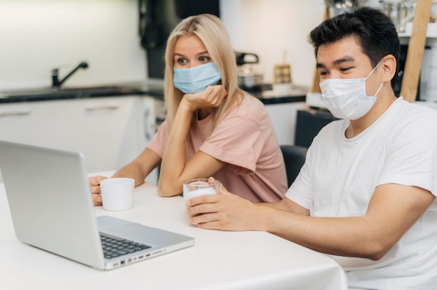 Pareja en casa con máscaras médicas durante la pandemia trabajando en una computadora portátil