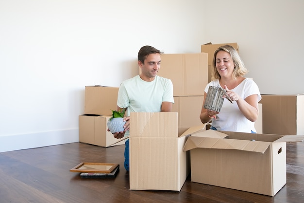 Pareja casada alegre que se muda a un nuevo apartamento, desembala cosas, se sienta en el suelo y saca objetos de cajas abiertas