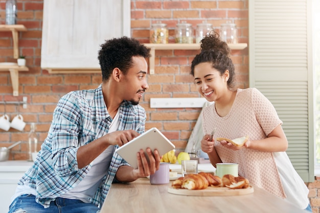 Pareja casada de raza mixta pasa el sábado por la mañana en la cocina, prepara bocadillos, usa una tableta