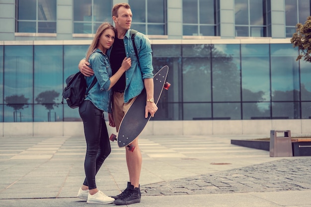 Pareja casual con longboard posando en una plaza de una ciudad.