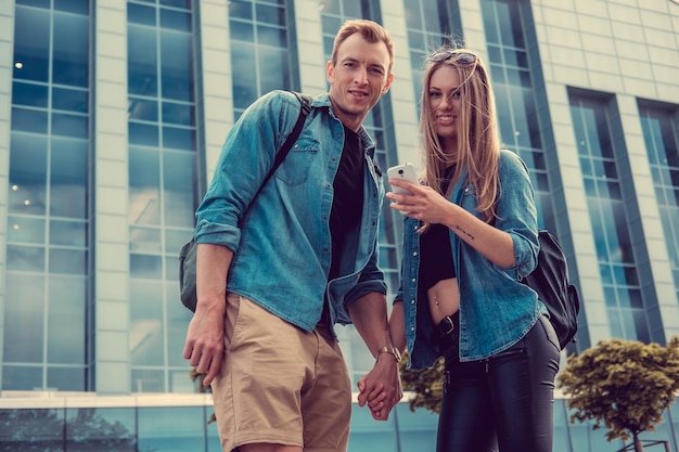 Pareja casual usando un teléfono inteligente y posando sobre un edificio de vidrio en el centro.