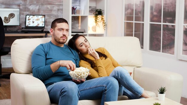 Pareja caucásica relajante viendo la televisión en la sala de estar comiendo palomitas de maíz y patatas fritas.