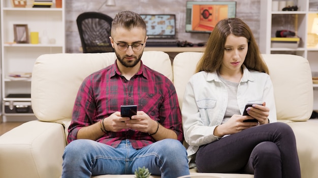 Pareja caucásica sentada en el sofá navegando en sus teléfonos. Novio y novia.