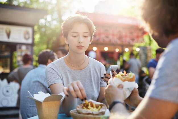 Pareja comiendo papas fritas y hamburguesas en un día soleado de verano en el parque en una feria pasando un buen rato.