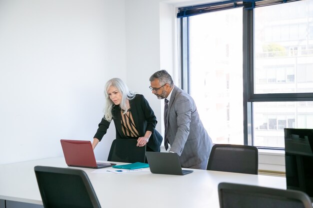 Pareja de compañeros de trabajo o socios que comienzan a reunirse en la sala de conferencias, de pie en la mesa y usan la computadora portátil juntos. Plano general. Concepto de comunicación empresarial