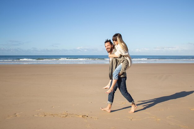 Pareja contenta pasando tiempo en la playa. Alegre mujer con ropa informal montando novios barbudos de vuelta. Amor, vacaciones, concepto de afecto.