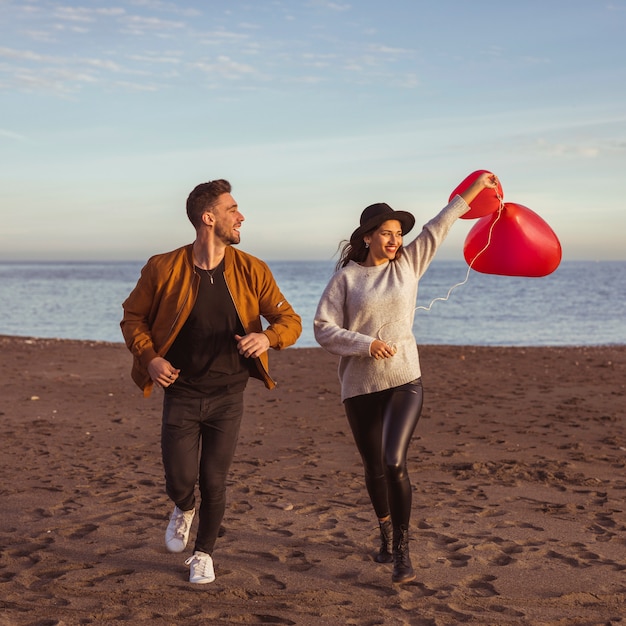 Foto gratuita pareja corriendo en la orilla del mar con globos de corazón