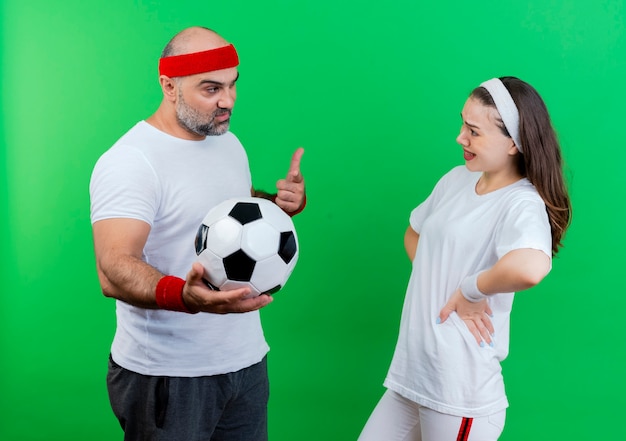 Pareja deportiva adulta con diadema y muñequeras hombre confiado sosteniendo y apuntando a un balón de fútbol mujer impresionada manteniendo las manos en la cintura mirándose aislado en la pared verde