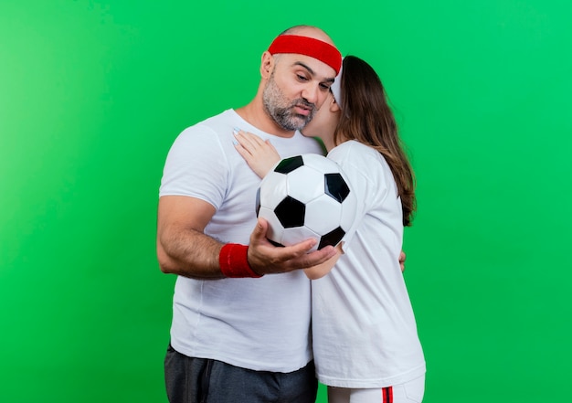 Pareja deportiva adulta vistiendo diadema y muñequeras abrazándose impresionado hombre sosteniendo y mirando una pelota de fútbol