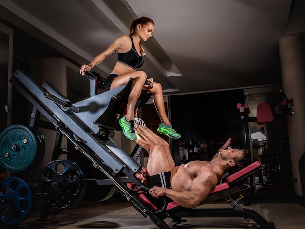 La pareja deportiva está haciendo ejercicio en el gimnasio. Hombre levantador de pesas haciendo prensas de piernas con su entrenador.