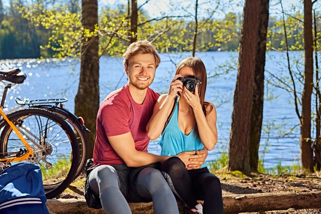 Una pareja deportiva positiva relajándose cerca del río después de un paseo en bicicleta. Mujer tomando fotografías en una cámara de fotos digital compacta.