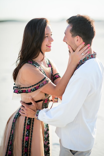 Foto gratuita pareja descalza en ropa bordada brillante abrazos tiernos en una arena blanca