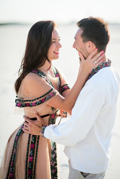 Pareja descalza en ropa bordada brillante abrazos tiernos en una arena blanca