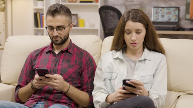 Pareja distante sentada en el sofá con sus teléfonos. Pareja navegando en sus teléfonos.