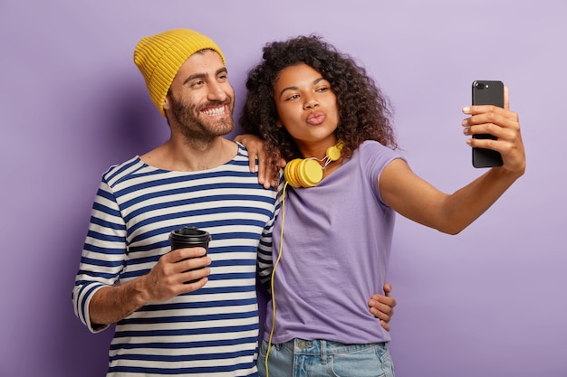 Pareja diversa positiva posa para hacer selfie, sonreír y hacer muecas del dispositivo, beber café para llevar, usar ropa informal, abrazarse contra la pared púrpura Tecnología, estilo de vida