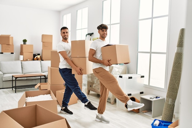 Pareja de dos hombres hispanos sonriendo confiados sosteniendo cajas de cartón en el nuevo hogar