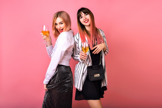 Pareja de dos mujeres elegantes bastante divertidas bebiendo champán y disfrutando de la fiesta, elegantes trajes blancos y negros de glamour, pelos rosados de moda