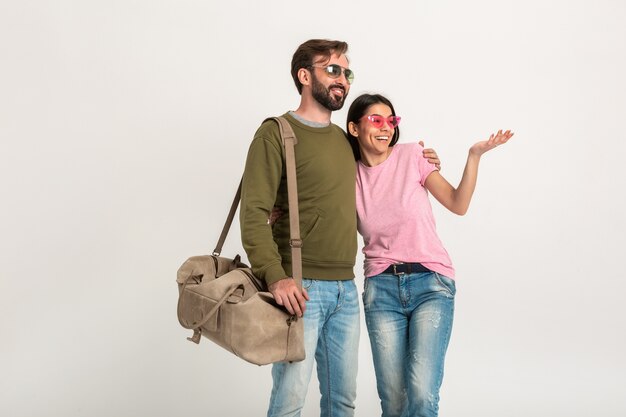 Pareja elegante aislada, mujer muy sonriente en camiseta rosa y hombre en sudadera con bolsa de viaje, vestido con jeans, gafas de sol, divirtiéndose juntos, señalando con el dedo