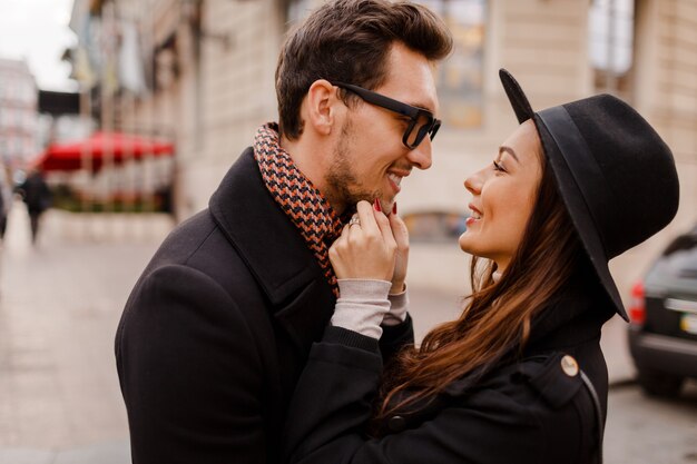 Pareja elegante de moda al aire libre en el amor vergonzoso y mirando el uno al otro con adoración. Mujer morena con gorro de lana con su novio en bufanda y abrigo.