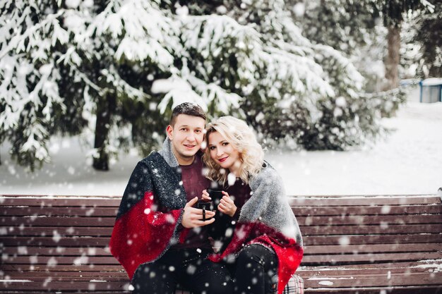 Pareja enamorada sentada en un banco y sonriendo en el bosque nevando.