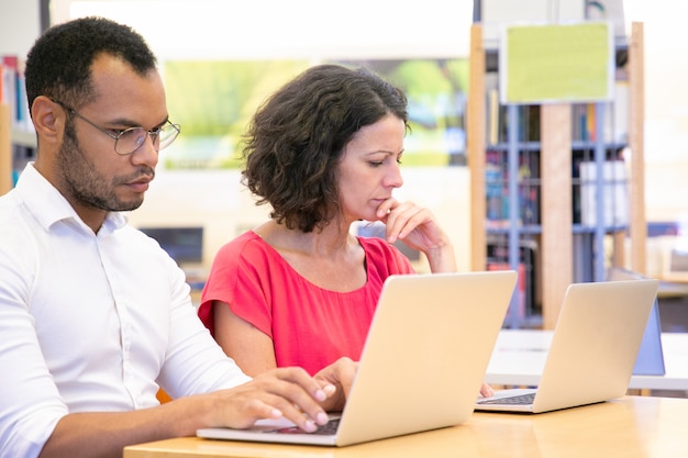 Pareja de estudiantes adultos serios trabajando en proyecto en biblioteca