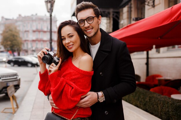 Pareja europea avergonzada y posando en la calle de vacaciones. Estado de ánimo romántico. Hermosa mujer morena con cámara de película.