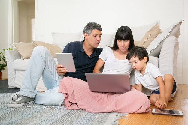 Pareja familiar con hijo pequeño usando computadoras portátiles, sentados en el piso del apartamento, disfrutando juntos del tiempo libre.