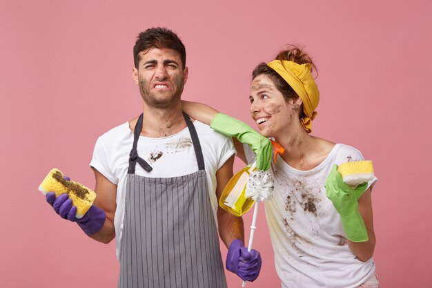 Pareja familiar ordenando su apartamento: hombre descontento con delantal sosteniendo una esponja sucia odiando la limpieza de pie cerca de su esposa que le pide que lave la ventana mirándolo con una sonrisa y amor