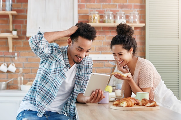 Pareja familiar positiva sonríe ampliamente mientras mira comedia en tableta, use conexión gratuita a Internet en casa,