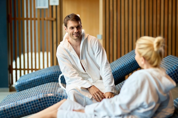 Pareja feliz en albornoces comunicándose mientras pasa un día relajante en el spa El foco está en el hombre