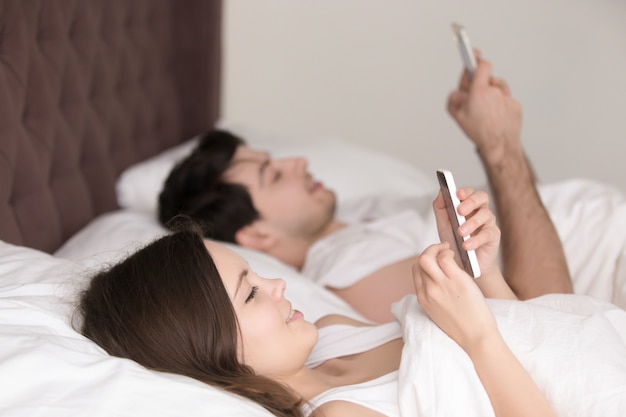 Pareja feliz cada uno usando su propio teléfono inteligente acostado en la cama