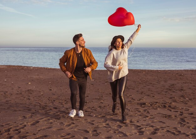 Pareja feliz corriendo en la orilla del mar con globos de corazón